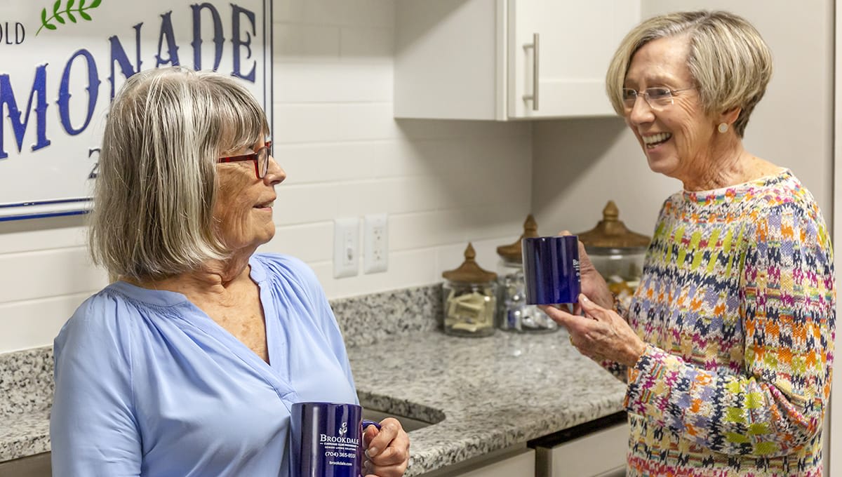 Two elderly women enjoying a conversation in the kitchen while holding mugs.