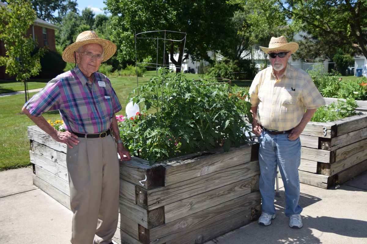 Two men in sun hats stand next to a raised garden bed filled with plants.