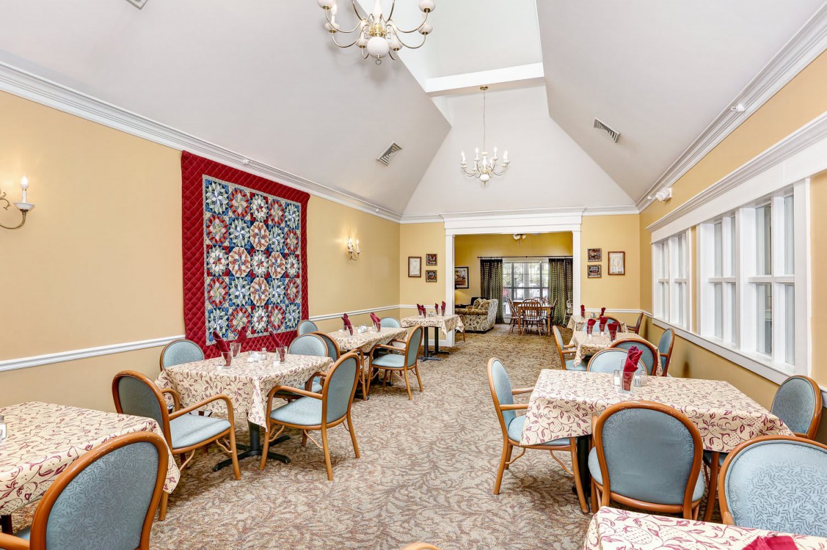 A cozy dining room with multiple tables and chairs, featuring a decorative quilt on the wall and a chandelier hanging from the ceiling.