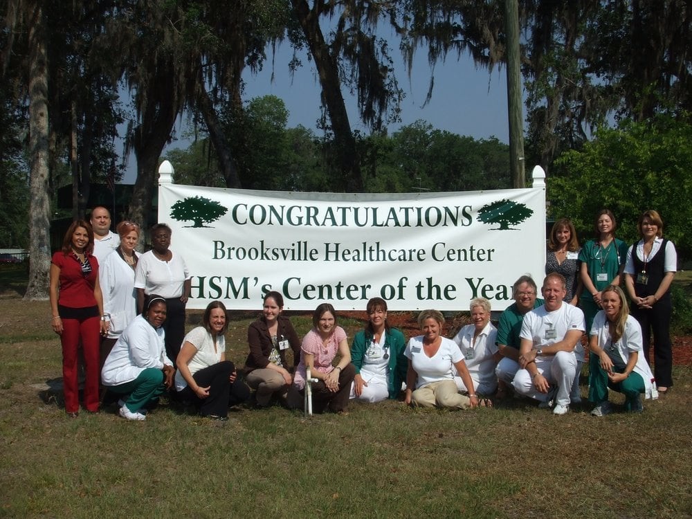 A group of people celebrating the Brooksville Healthcare Center being named HSM's Center of the Year, outdoors with a congratulatory banner.