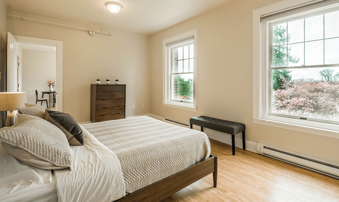 A minimalist bedroom with neutral colors, featuring a bed, dresser, and ample natural light from two windows.