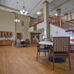 A spacious dining area with wooden flooring, featuring tables and chairs under high ceilings adorned with lighting fixtures.