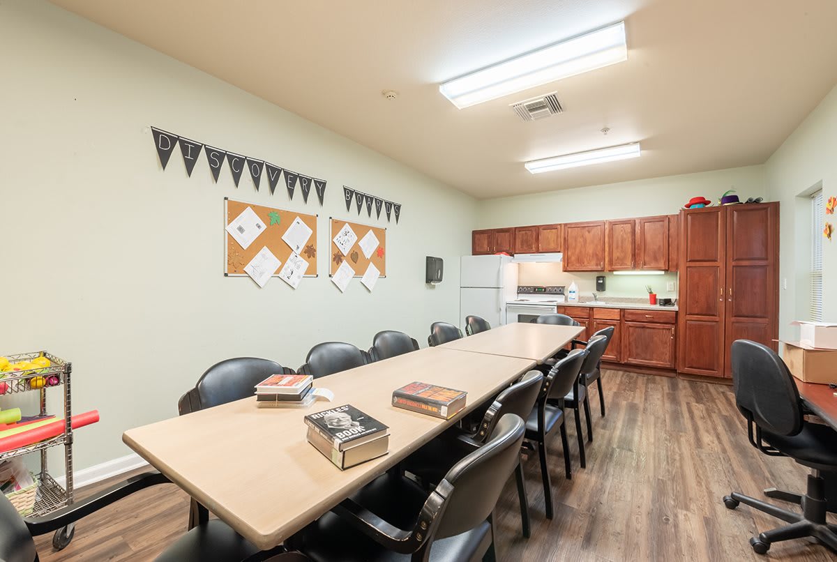 A well-lit conference room with a long meeting table, surrounded by chairs and a kitchenette at the back.