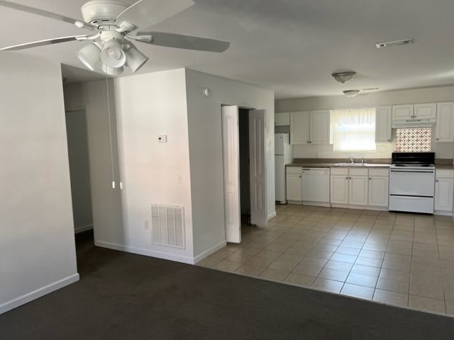 A view of a living room and kitchen area in an open-plan layout, featuring a ceiling fan and white cabinetry.