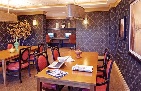 A well-decorated dining room featuring a wooden table with chairs, a laptop, and books. In the background, there are computer workstations against a wall with patterned wallpaper and elegant lighting fixtures.