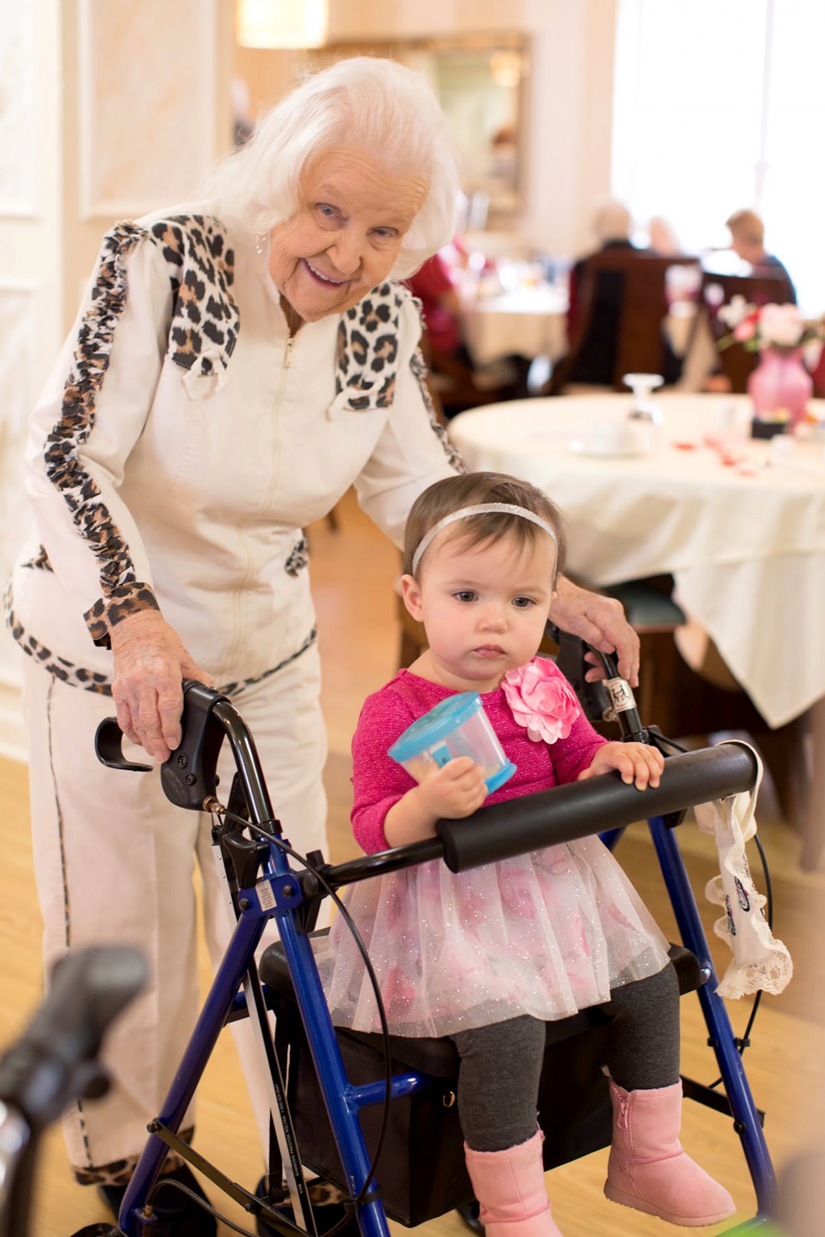 An elderly woman assists a toddler with a walker during a family event.