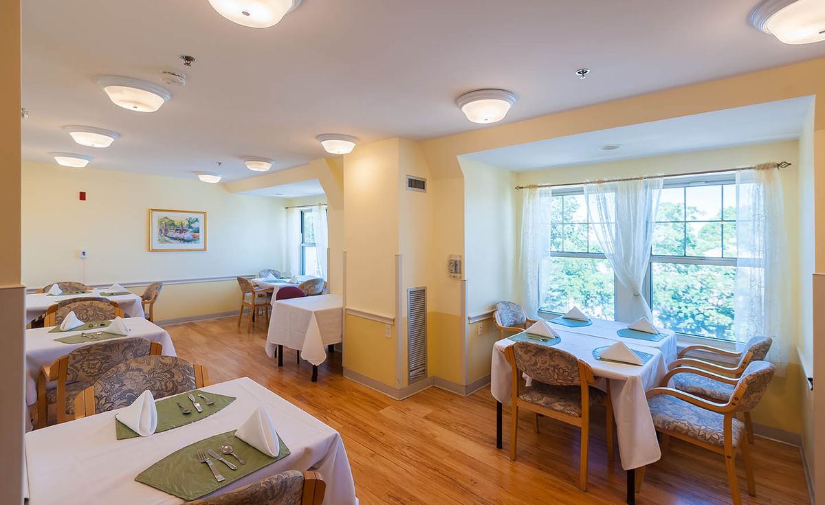 A brightly lit dining room with neatly arranged tables and chairs, featuring large windows that allow natural light to fill the space.