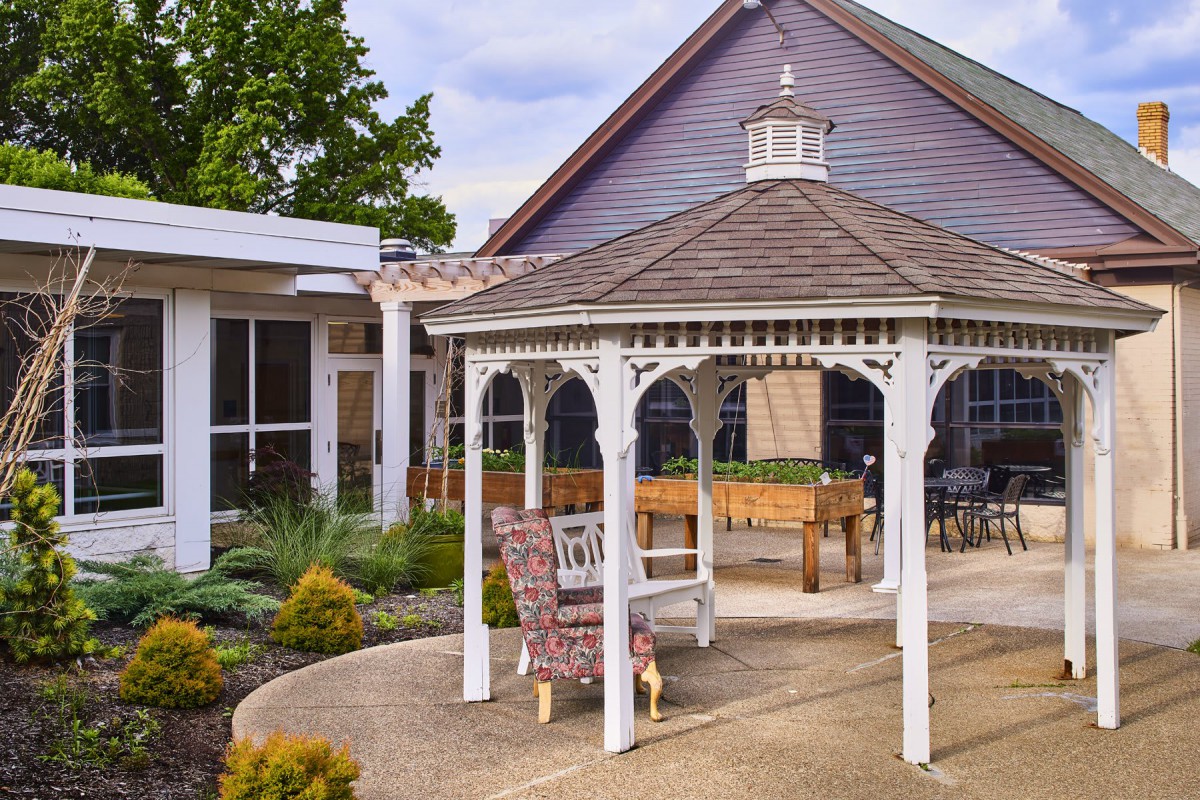 A peaceful garden setting with a white gazebo and comfortable seating, surrounded by landscaped greenery and a charming house.