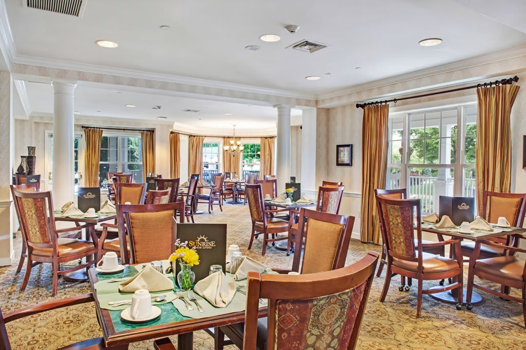 A spacious dining room with elegantly set tables and wooden chairs, featuring large windows and soft lighting.