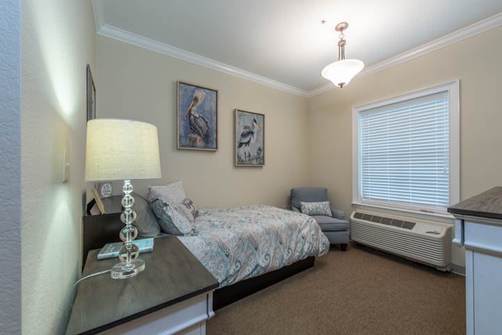 A cozy bedroom with a bed, chair, and wall art, softly lit by a lamp and natural light from the window.