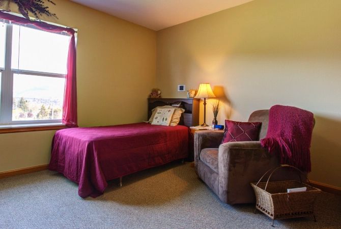 A cozy bedroom featuring a single bed with a maroon bedspread, an armchair beside a small lamp on a nightstand, and a window providing natural light.