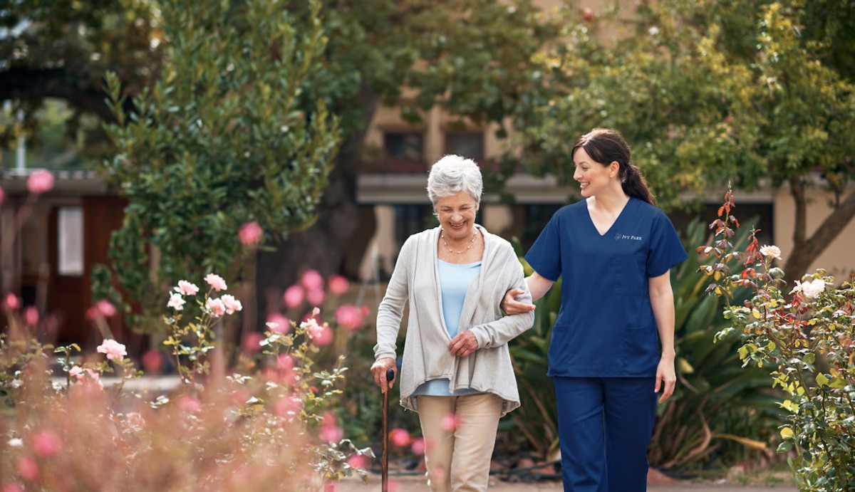 A nurse walking alongside an elderly woman in a garden.