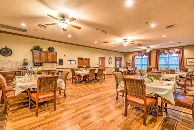 A spacious dining area with wooden floors, neatly arranged tables and chairs, ceiling fans, and warm lighting.