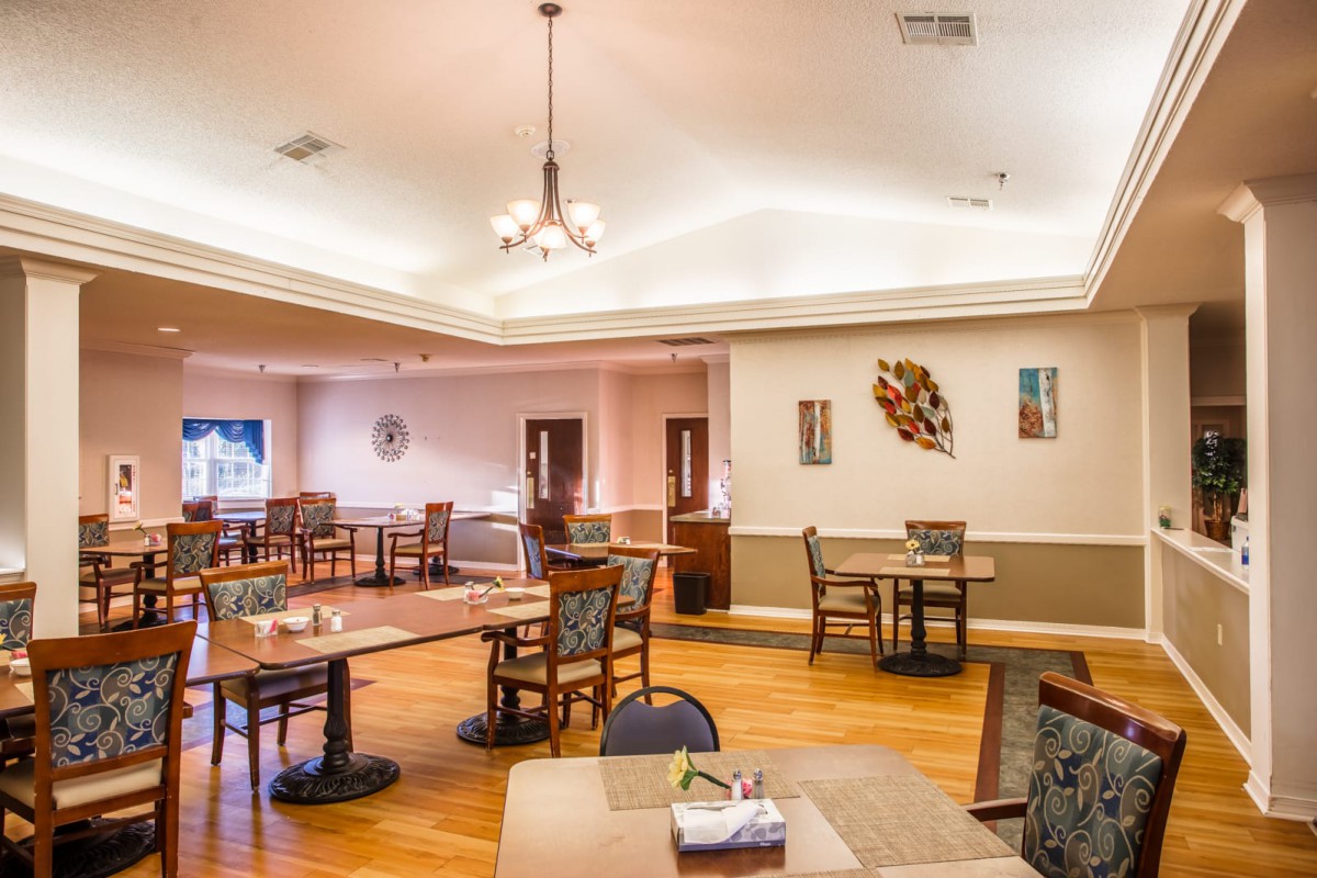A spacious dining room with wooden flooring, featuring multiple tables and chairs, adorned with a chandelier and artistic wall decorations.