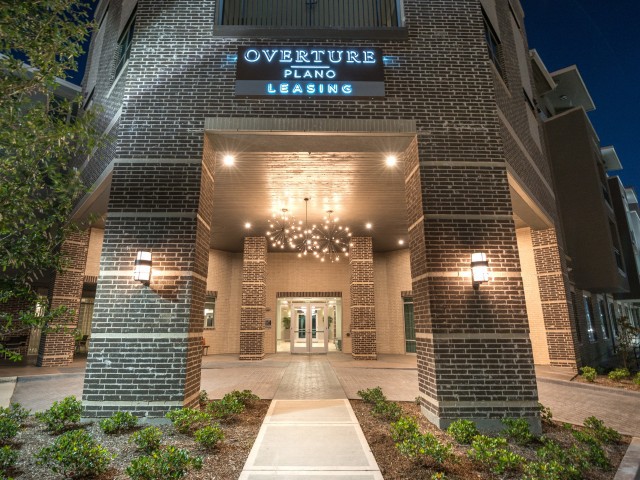 The illuminated entrance of Overture Plano at night, featuring modern architecture and a prominent chandelier.