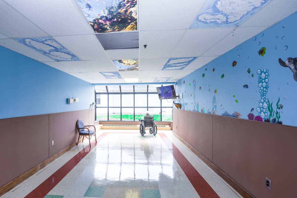 A hospital corridor with colorful ceiling art and a person in a wheelchair near a large window.