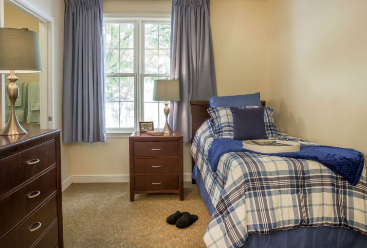 A cozy bedroom with a single bed featuring plaid bedding, a nightstand with a lamp, and curtains framing a window.