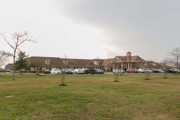 A large building with a parking lot in front, surrounded by a lawn and trees under a cloudy sky.