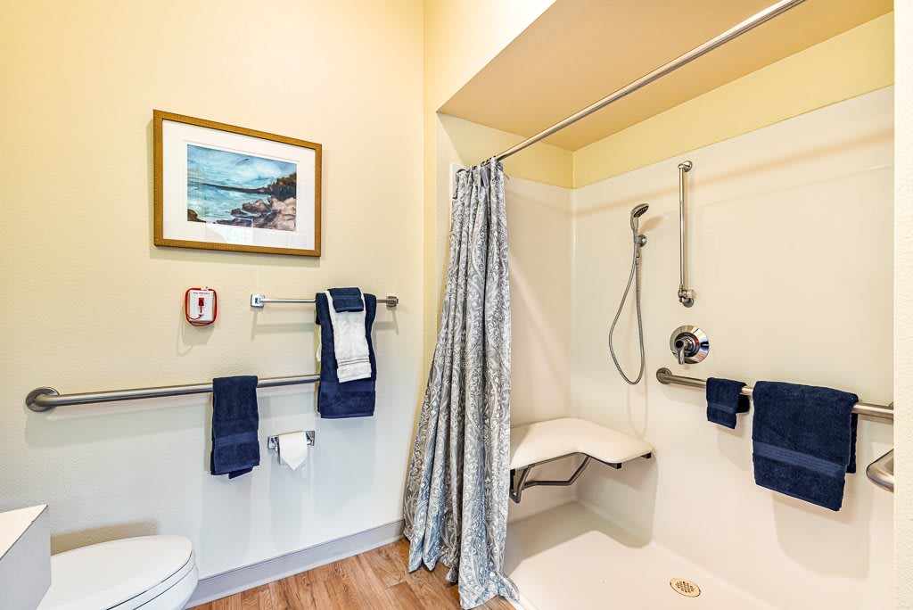An accessible bathroom with a shower featuring handrails, a foldable chair, and towel racks.