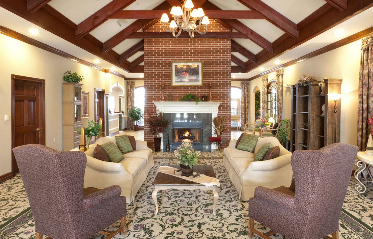 A luxurious living room featuring a central fireplace, elegant sofas, armchairs, and a decorative rug under a vaulted ceiling with an ornate chandelier.