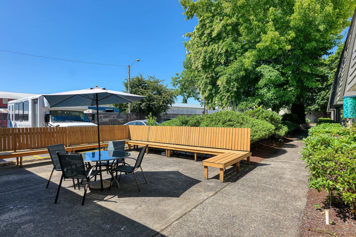 A sunny outdoor patio with a table, chairs, and benches under an umbrella surrounded by greenery.