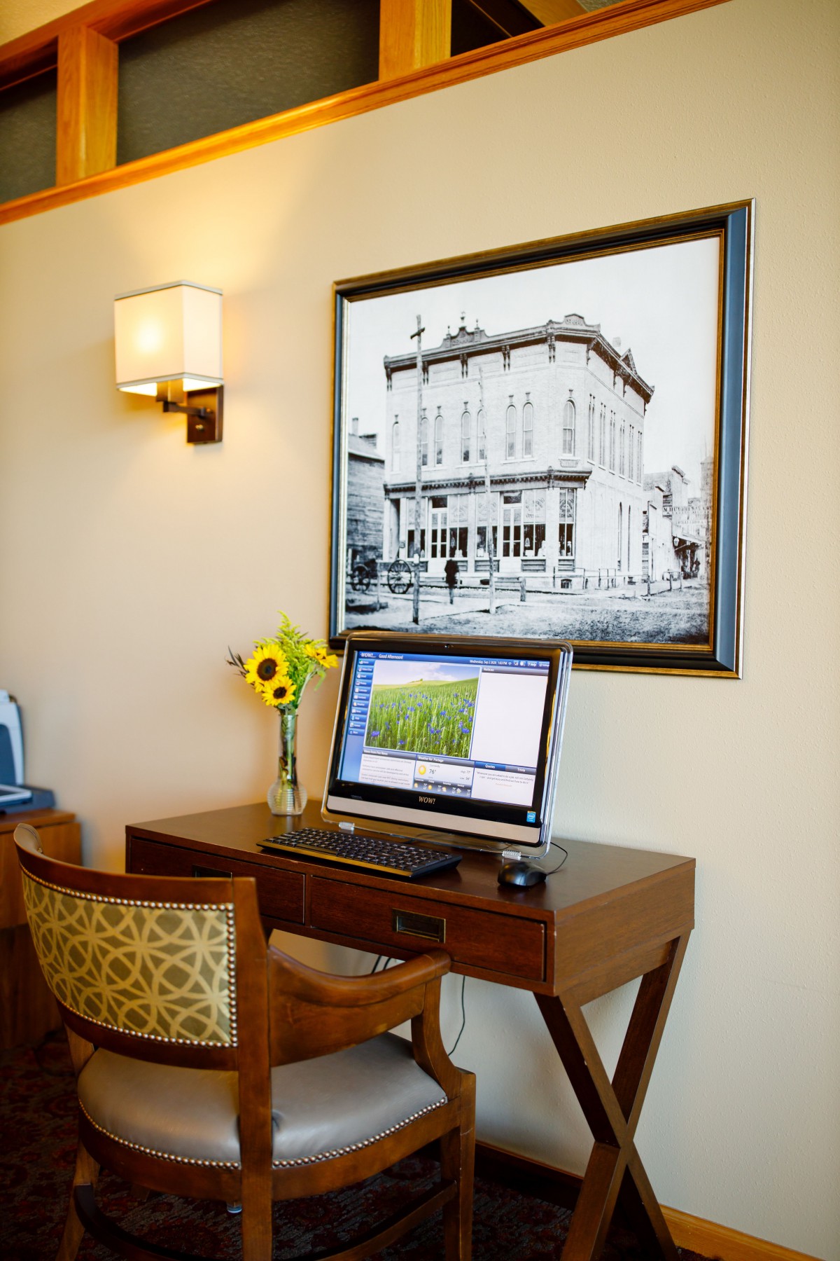 A cozy office setup with a computer on a wooden desk, adorned with flowers and illuminated by a wall lamp.