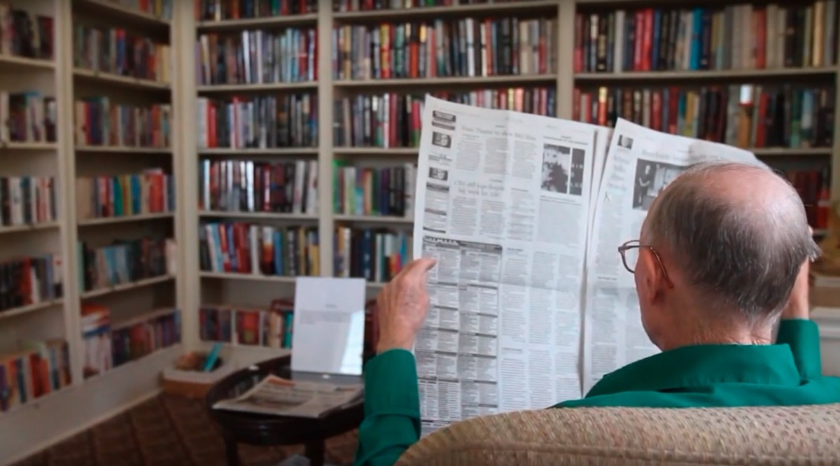 A person sitting in a library, reading a newspaper with bookshelves in the background.