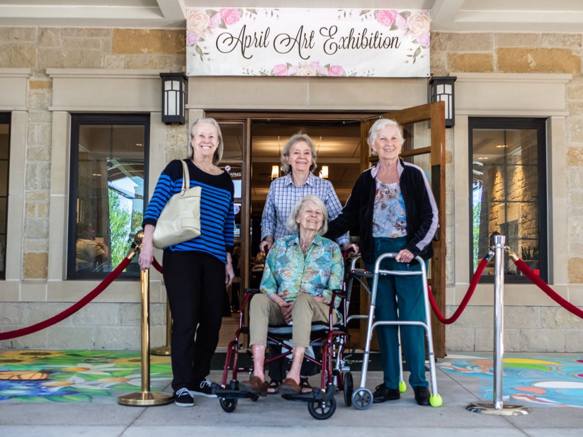 A group of elderly women pose in front of an art exhibition entrance, with one using a wheelchair and another a walker.