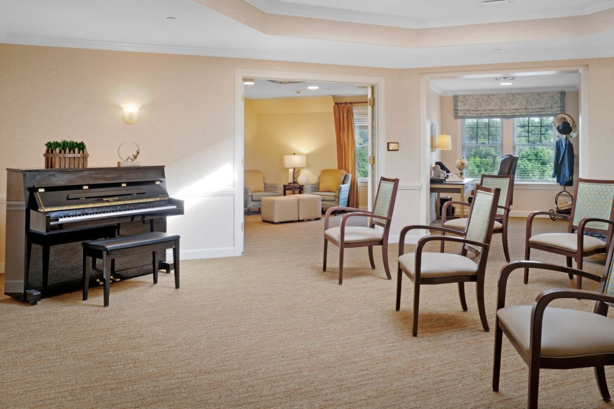 A spacious, carpeted room with a piano and several chairs, leading into a cozy living area.