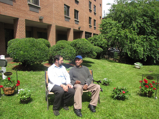 Two individuals engaged in a conversation on a bench in a grassy area surrounded by bushes.