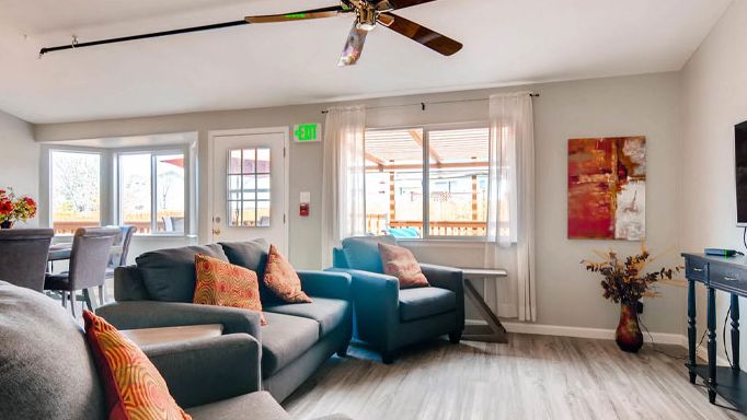 A cozy living room with comfortable seating and a ceiling fan, illuminated by natural light from the window.