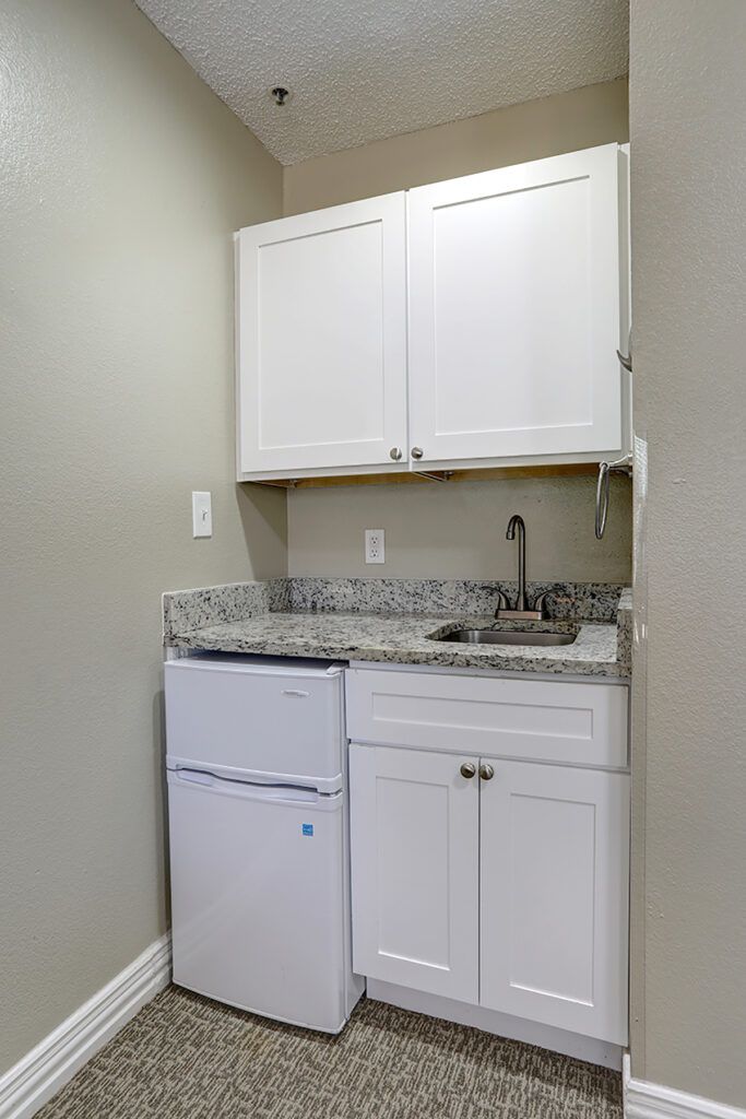 Compact kitchenette area with white cabinets, granite countertop, and a small refrigerator.