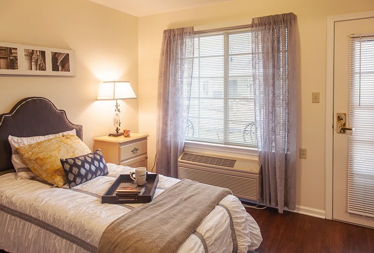 A cozy bedroom with a neatly made bed, soft lighting, and a window with sheer curtains.