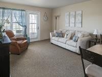 A cozy living room with a sofa and armchair, carpeted floor, and large window with sheer curtains.