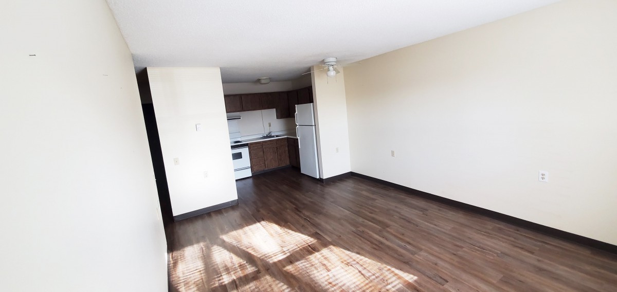 An empty apartment with a hardwood floor, featuring a small kitchen area and sunlight streaming through the windows.