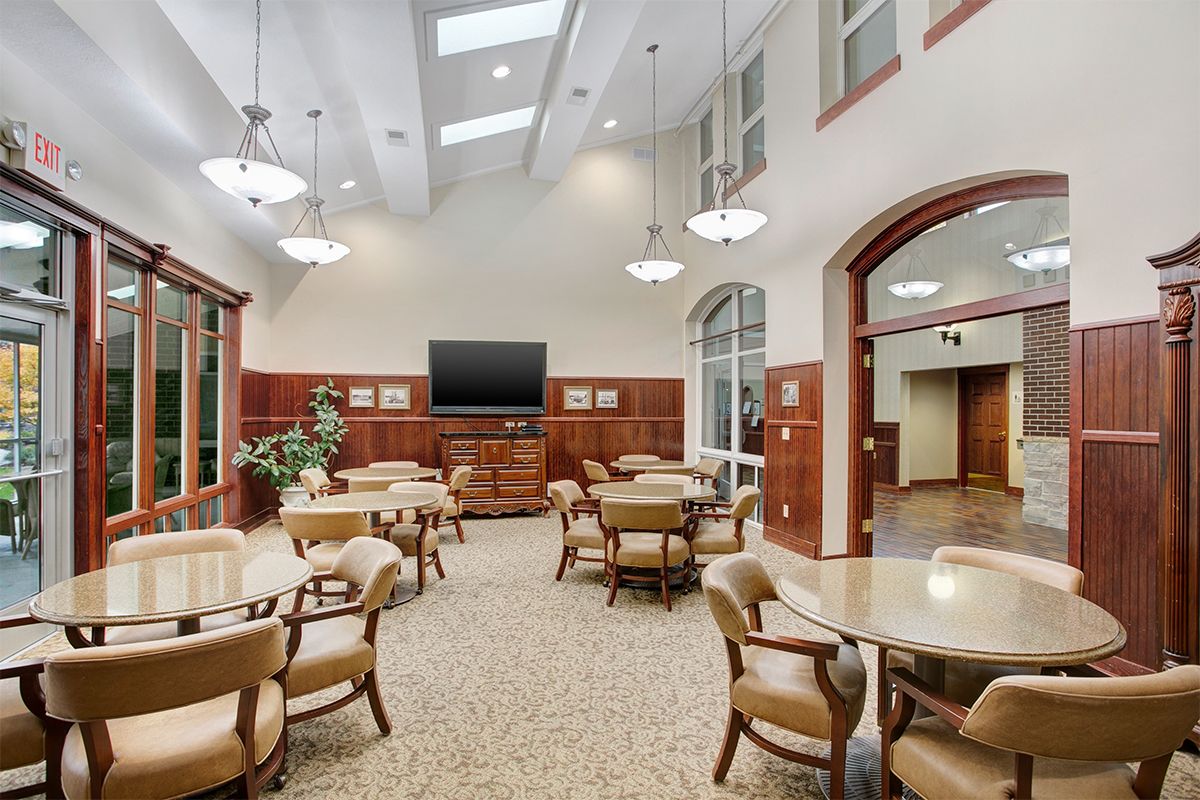 A spacious and well-lit community room featuring multiple tables and chairs, a large television, and skylights.