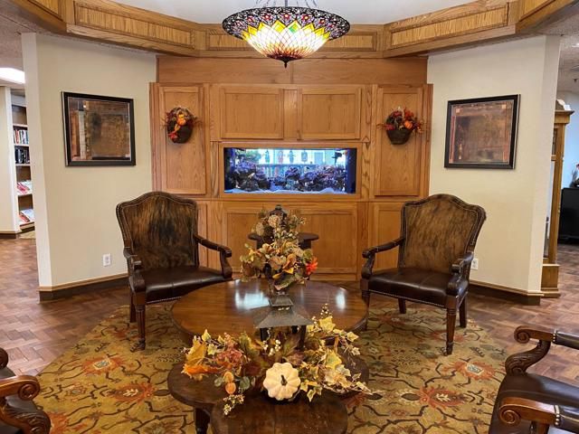 A cozy sitting area featuring two wooden chairs, a central table with floral arrangements, and an aquarium set in wood-paneled walls.