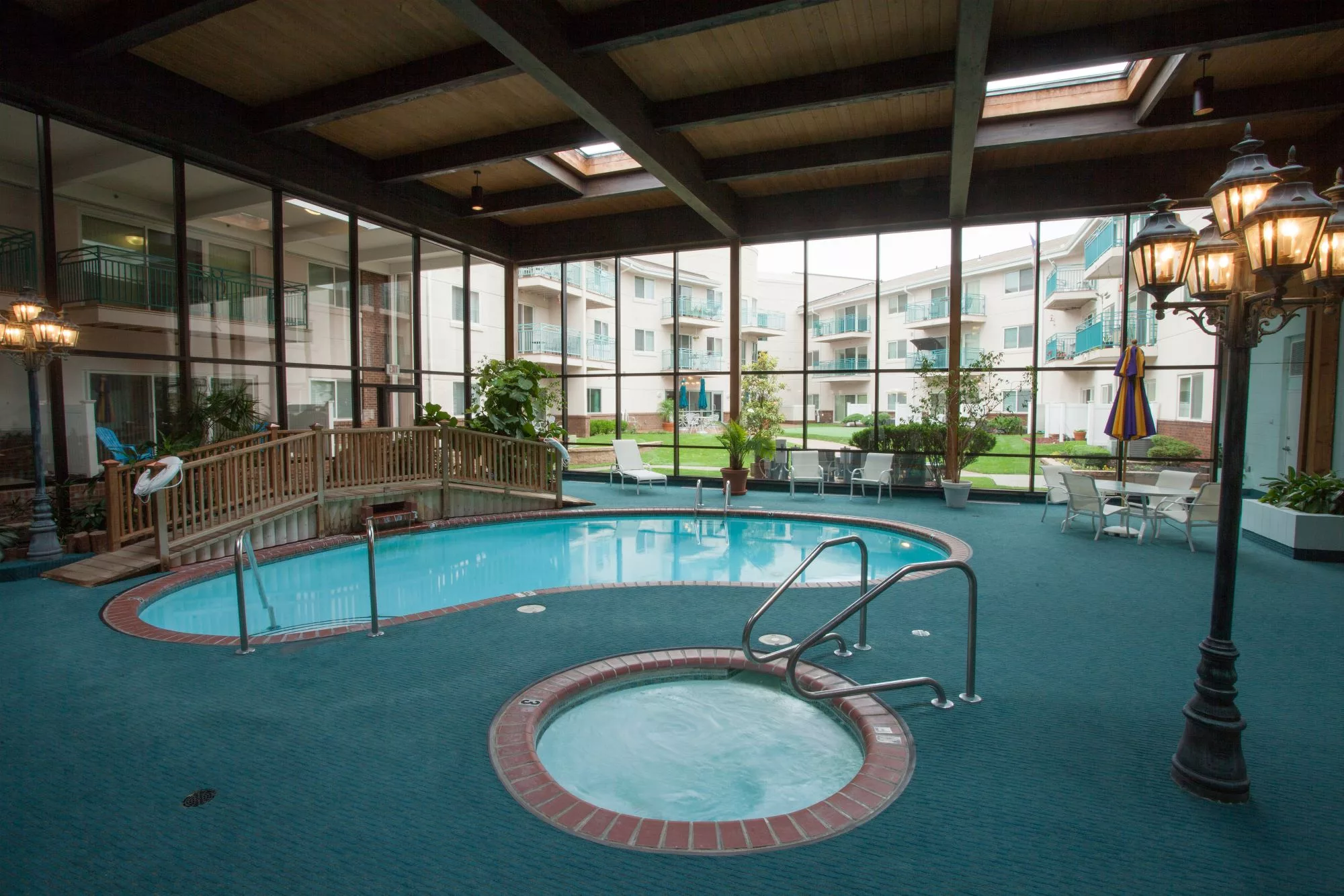 An indoor swimming pool area featuring a hot tub, surrounded by large glass windows with views of an outdoor complex.