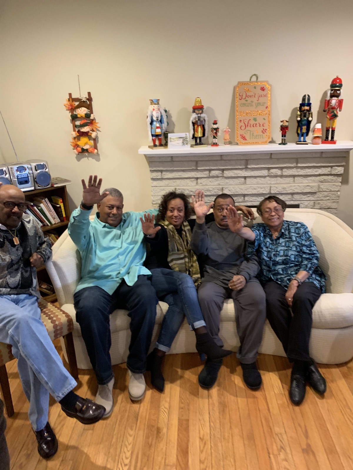 A group of people sitting on a couch in a cozy living room, smiling and waving at the camera.