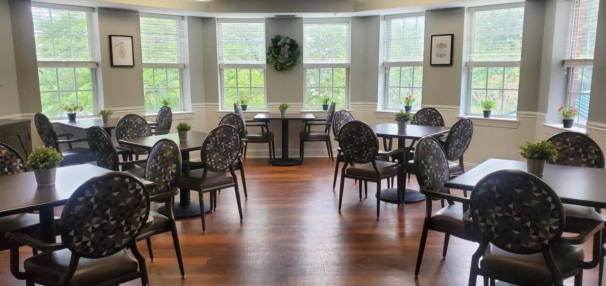 A cozy cafe interior with round tables and decorative chairs, illuminated by natural light from large windows.