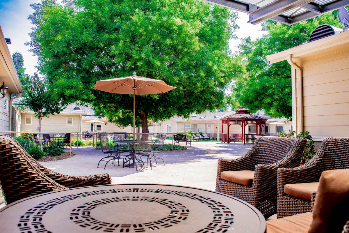 A serene outdoor patio area featuring wicker chairs and a table under an umbrella, with lush trees and a gazebo in the background.