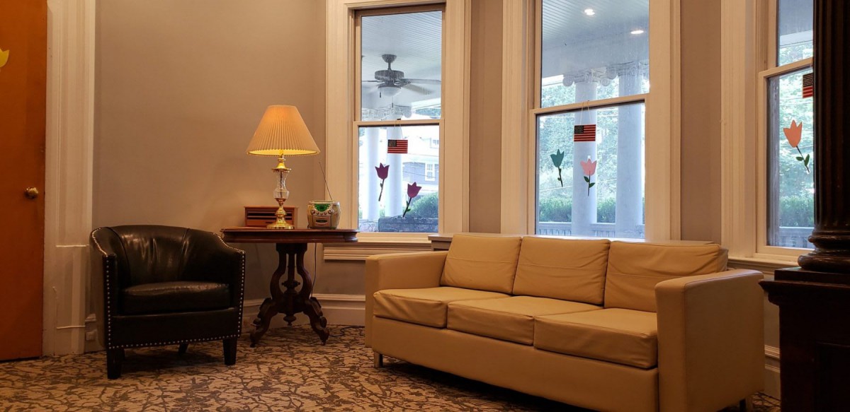 A cozy living room featuring a beige sofa, a side table with a lamp, and large windows overlooking a porch.