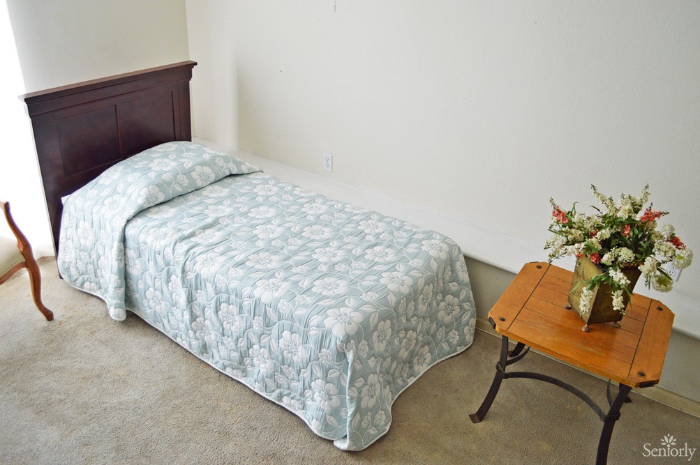 A simple bedroom with a single bed, covered in a patterned blanket, and a small table beside it holding a vase with flowers.