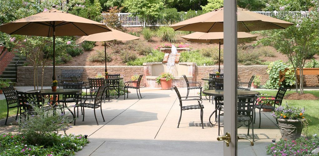 A serene outdoor patio area with tables, chairs, and umbrellas, featuring a central fountain surrounded by lush greenery.
