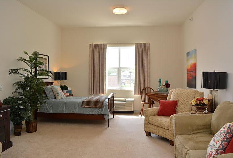 A cozy bedroom with a large window, featuring a bed, armchair, and various decorative elements.