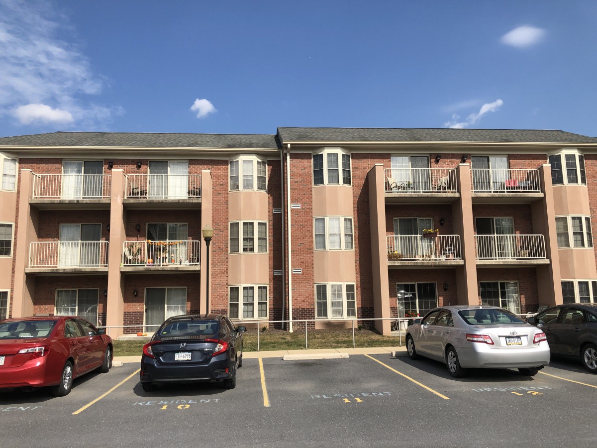A three-story brick apartment building with balconies and parked cars in front under a clear sky.