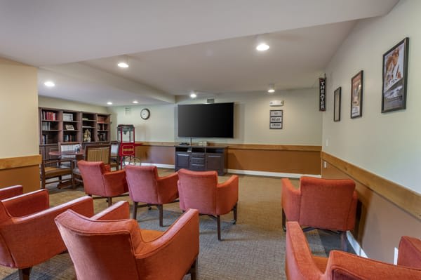 A conference room with multiple chairs facing a large television, featuring a bookshelf and carpeted flooring.