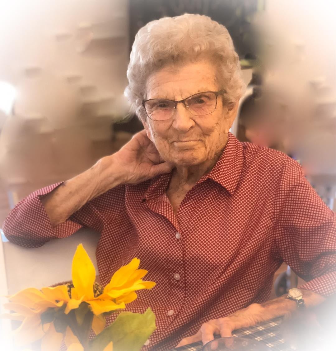 An elderly woman wearing glasses and a red shirt, sitting indoors with yellow flowers in the foreground.