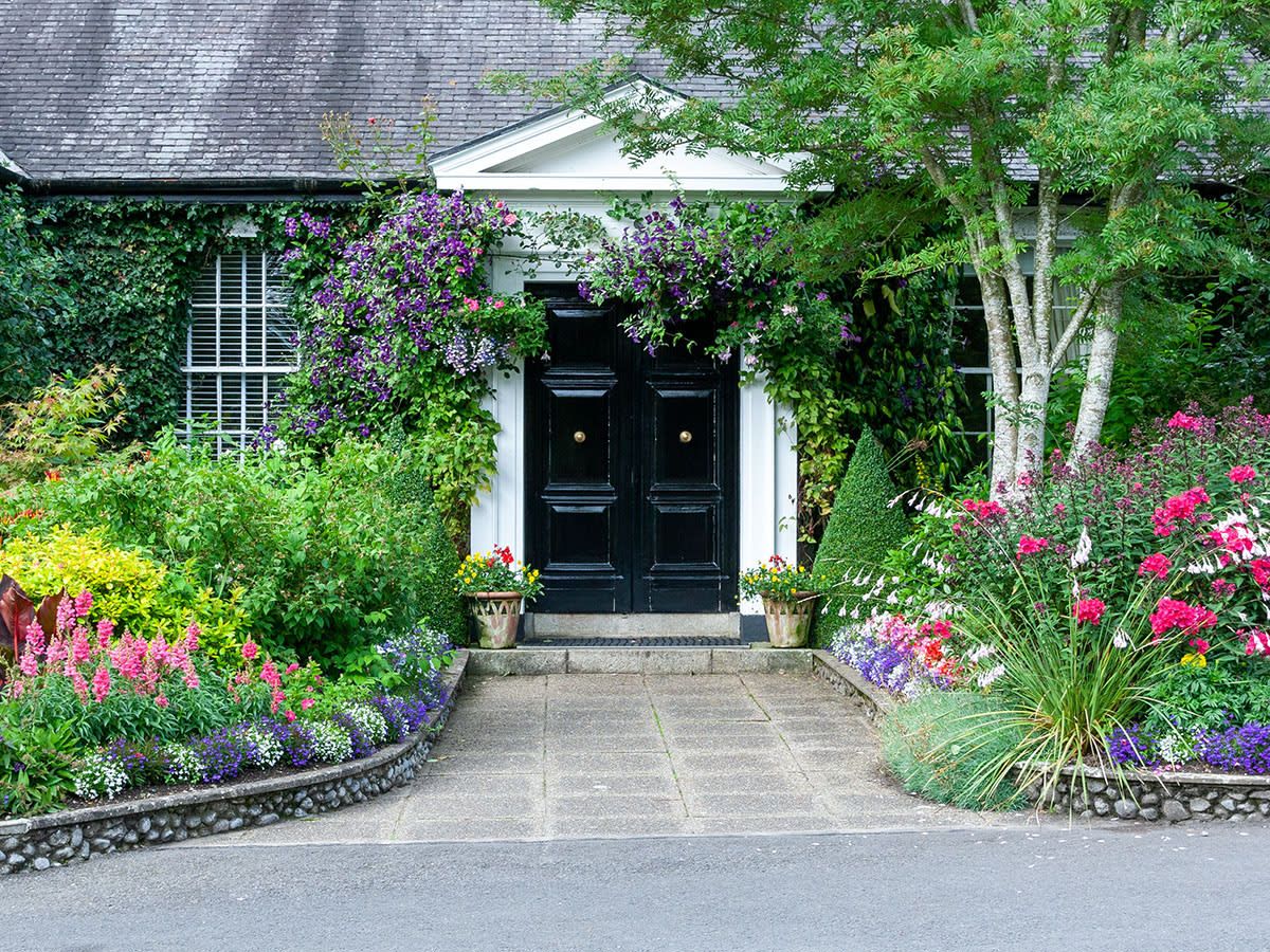 A charming garden path leads to a house with a black double door, surrounded by colorful flowers and lush greenery.