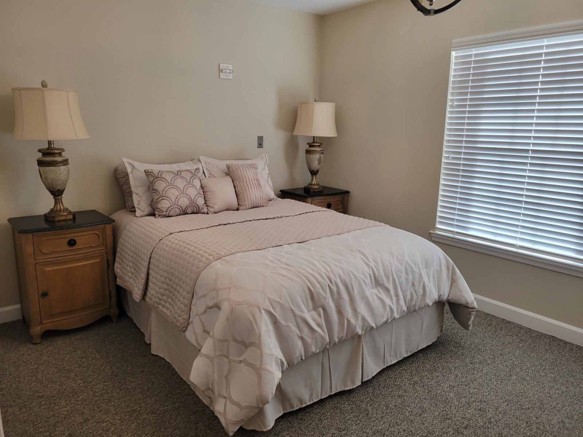 A cozy bedroom featuring a neatly made bed with decorative pillows, flanked by nightstands and lamps, and a window with blinds.